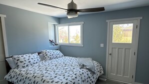 Un dormitorio con una cama, ventilador de techo, dos ventanas y una puerta.