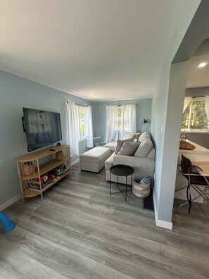 Un salon avec un canapé gris, un téléviseur posé sur un meuble, une étagère en bois et un coin repas comprenant une table et des chaises.