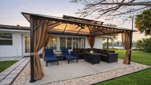 En udendørs patio med pergola, siddeområde og udsigt til et moderne hus.