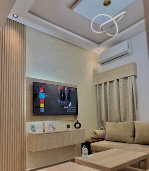 A modern living room with a flat-screen TV mounted on a wooden wall, a ceiling light fixture, and a sofa with a cushion. - Atlas 001 (Tangier)