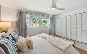 Een slaapkamer met een bed, gordijnen, een plafondventilator en uitzicht op een tuin door het raam.