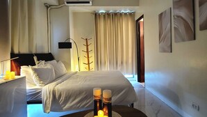 A modern hotel room with a bed, bedside table, lamp, and a small table with candles. - Spectacular 2.5BR Connected to Eastwood  Mall (Quezon City)