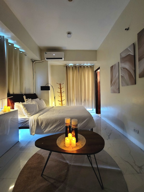 A modern hotel room with a bed, bedside table, lamp, and a small table with candles.