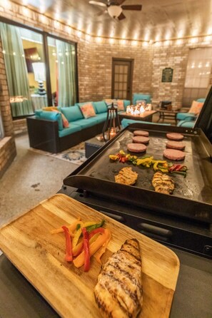 A grill with food, a wooden cutting board with vegetables, and a seating area with sofas and a table.
