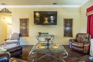 A living room with a flat-screen TV mounted on the wall, a glass coffee table with two cups and a bowl, a brown leather sofa, and a beige armchair.
