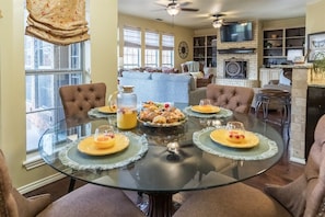 A dining area with a glass table set for six, surrounded by chairs, with a kitchen and living area in the background. - The Regent Abilene Luxury Retreat w Patio Fire (Abilene)
