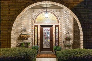 A brick building entrance with a wooden door, a welcome mat, and two flower baskets on either side. - The Regent Abilene Luxury Retreat w Patio Fire (Abilene)