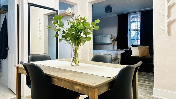 A dining area with a wooden table and black chairs, a vase with green foliage, and a view into a living room with a sofa and a window. - Hidden Gem in the Heart of Capitol Hill (Denver)