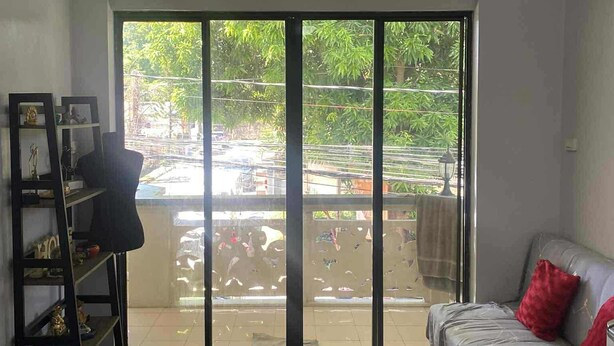 Ein Zimmer mit Deckenventilator, einer Couch mit rotem Kissen, Holzboden und einem großen Fenster mit Blick auf Grünflächen und einen Parkplatz.