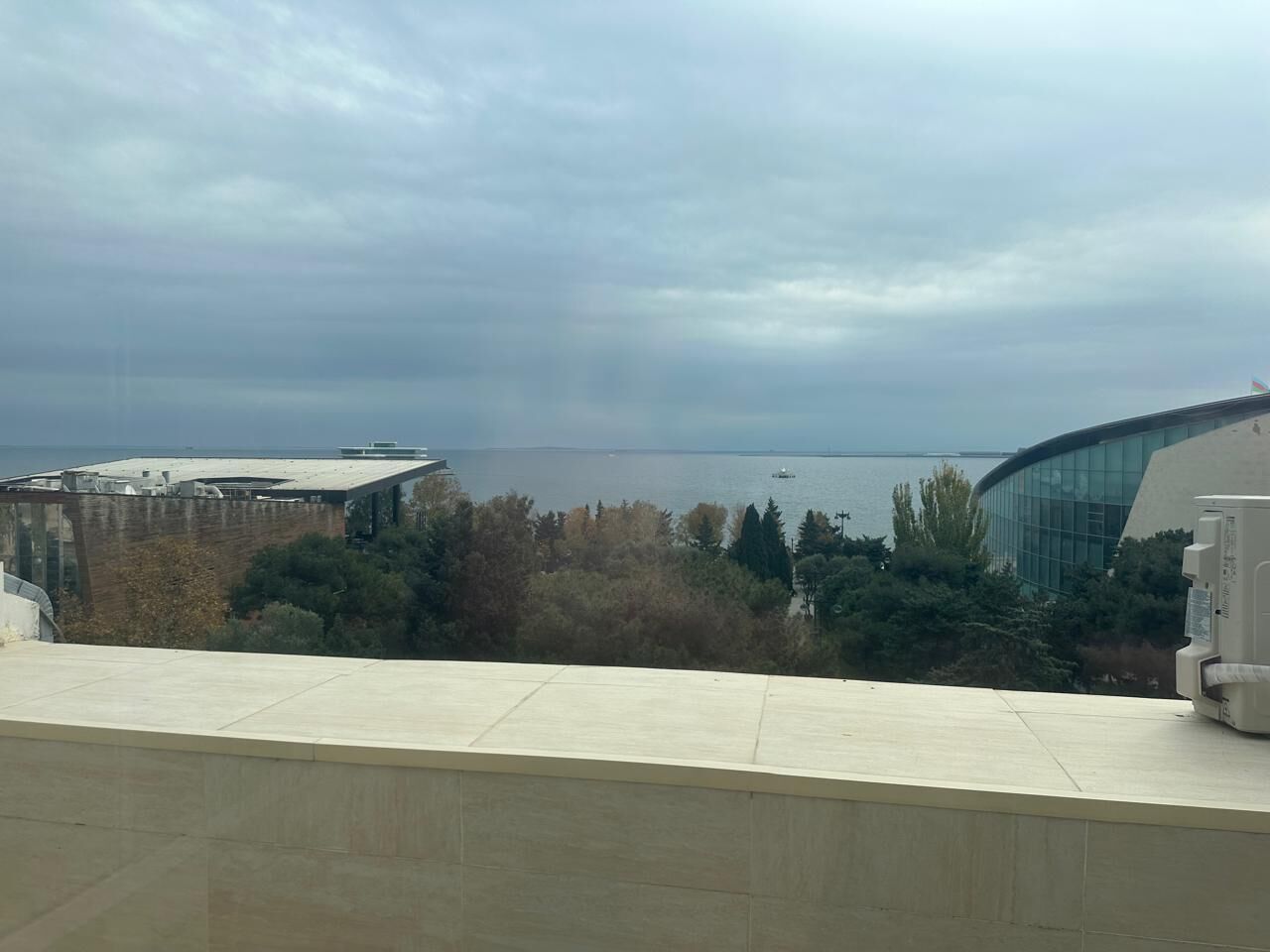 Blick auf ein modernes Gebäude mit einer geschwungenen Glasfassade, dahinter ein Gewässer und ein bewölkter Himmel.
