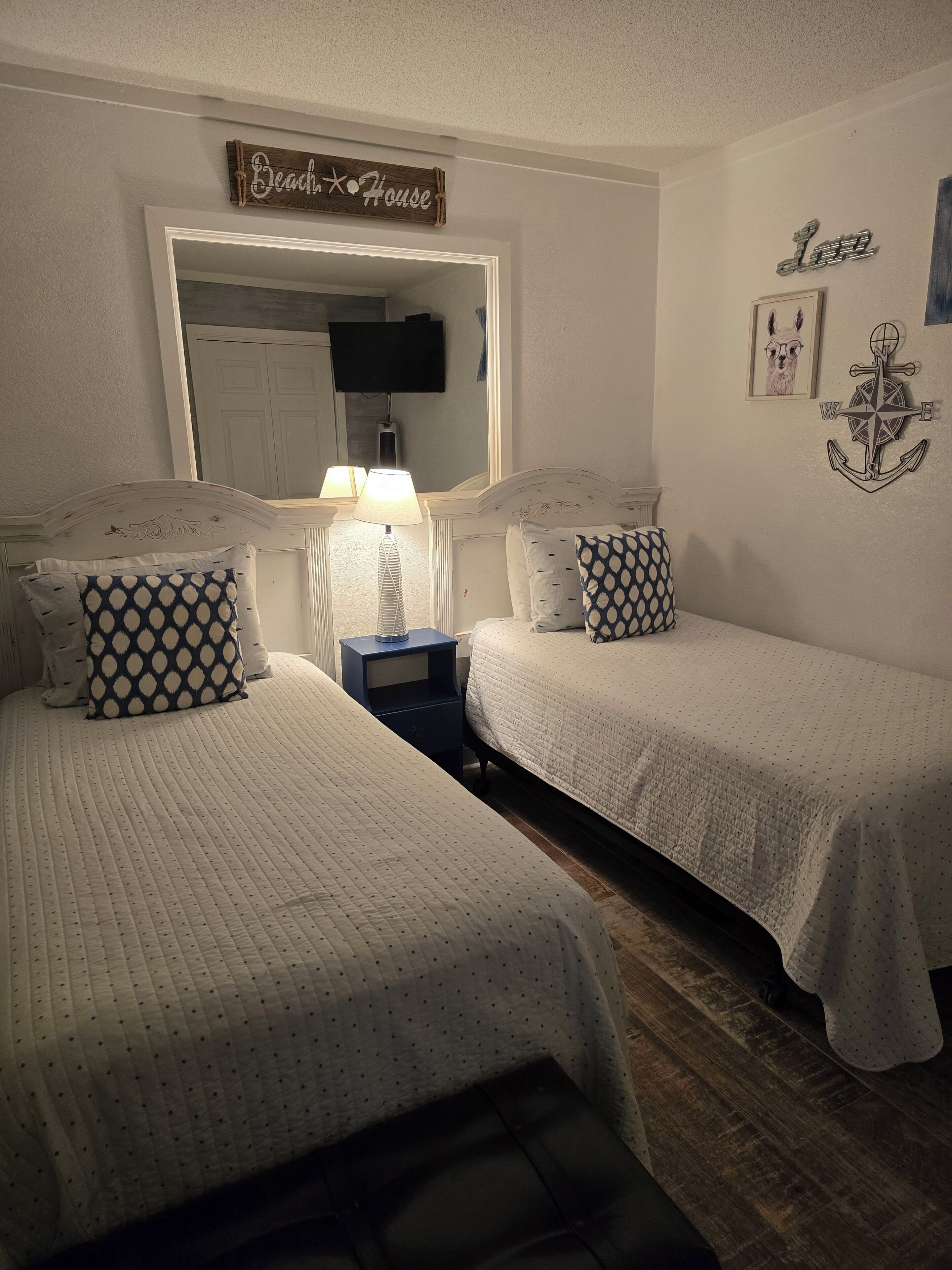 A bedroom with two beds, a nightstand, a lamp, a mirror, and wall decorations including a compass and a sign that reads 'Beach House'.