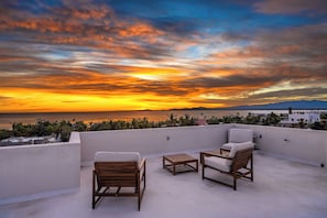 A rooftop terrace with two wooden chairs and a small table, overlooking a sunset over the sea with palm trees and buildings in the background.