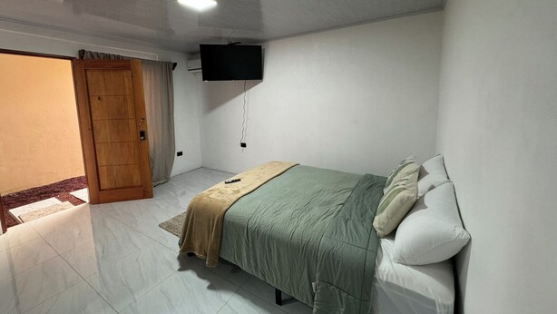 A bedroom with a bed, a TV mounted on the wall, a wooden door, and a tiled floor. - Walk Everywhere , Budget Stay w/ AC + WiFi #1 (La Fortuna)
