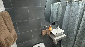 A bathroom with dark tiles, a white toilet, a white sink, a mirror, and a shower curtain. - Walk Everywhere , Budget Stay w/ AC + WiFi #1 (La Fortuna)