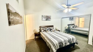 Ein Schlafzimmer mit einem Bett, einem Nachttisch, einer Lampe, einem Deckenventilator und einem Spiegel.