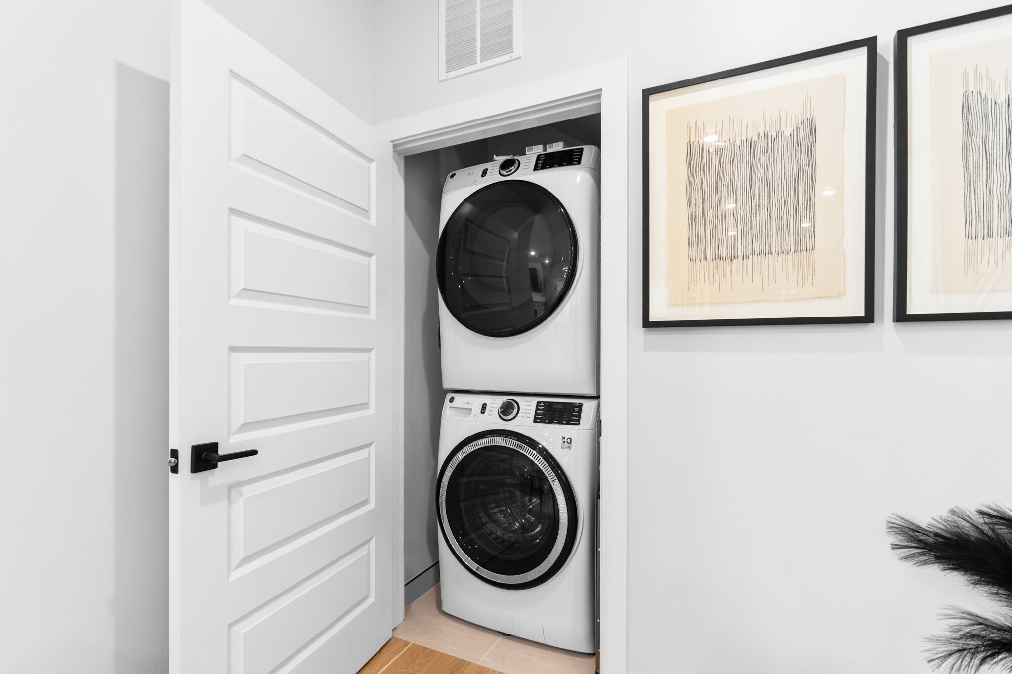 A laundry room with a front-loading washer and dryer, a white door, and framed artwork on the wall.