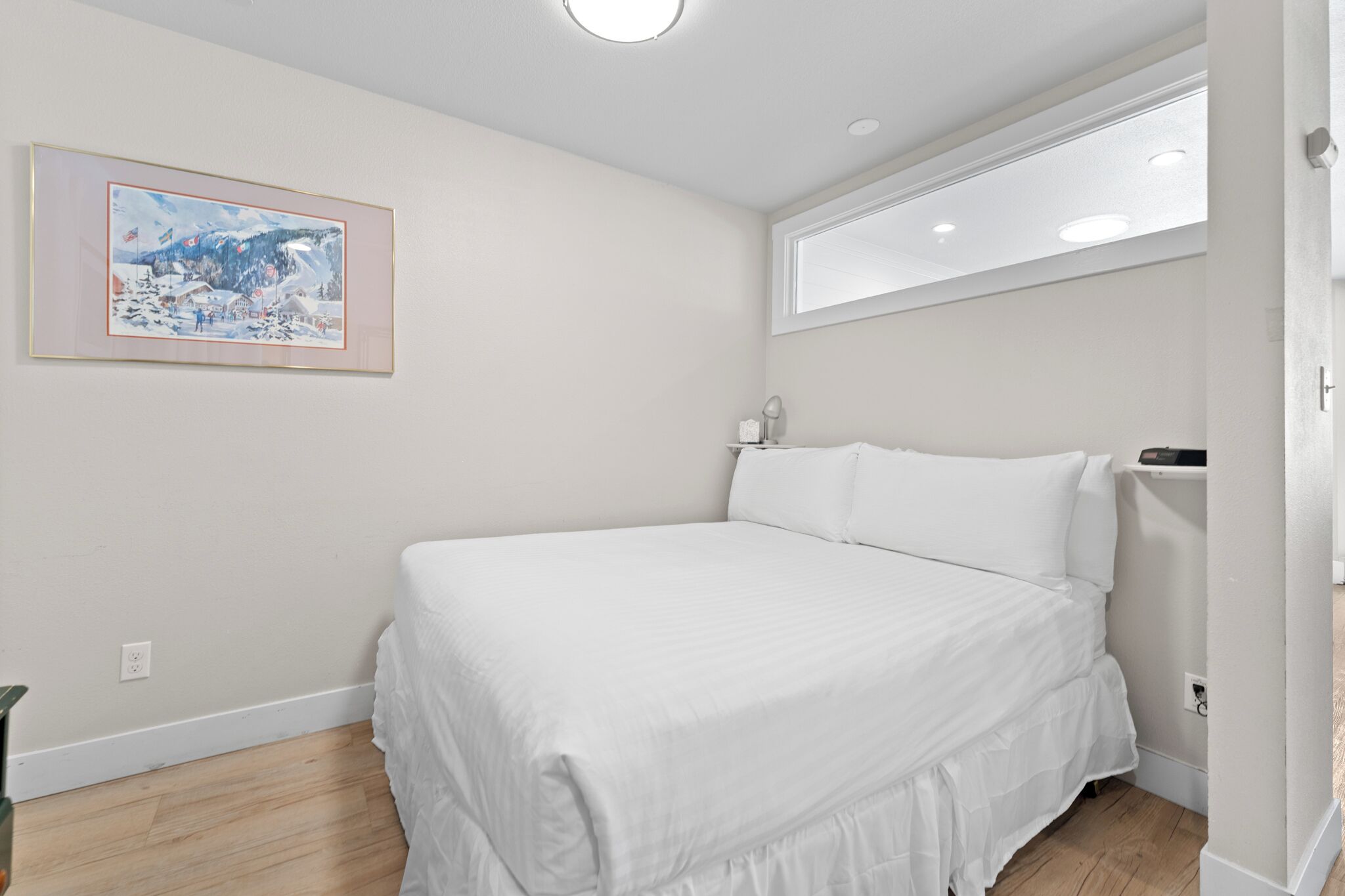 A bedroom with a large bed, a window, a painting on the wall, and a ceiling light.