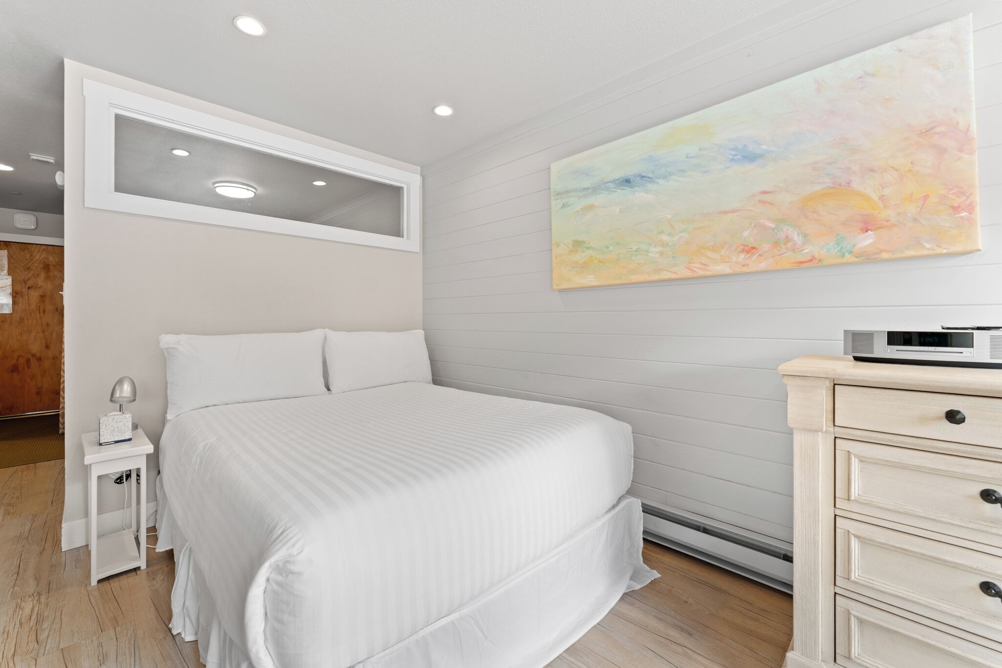 A bedroom with a bed, bedside table, lamp, dresser, and a painting on the wall.