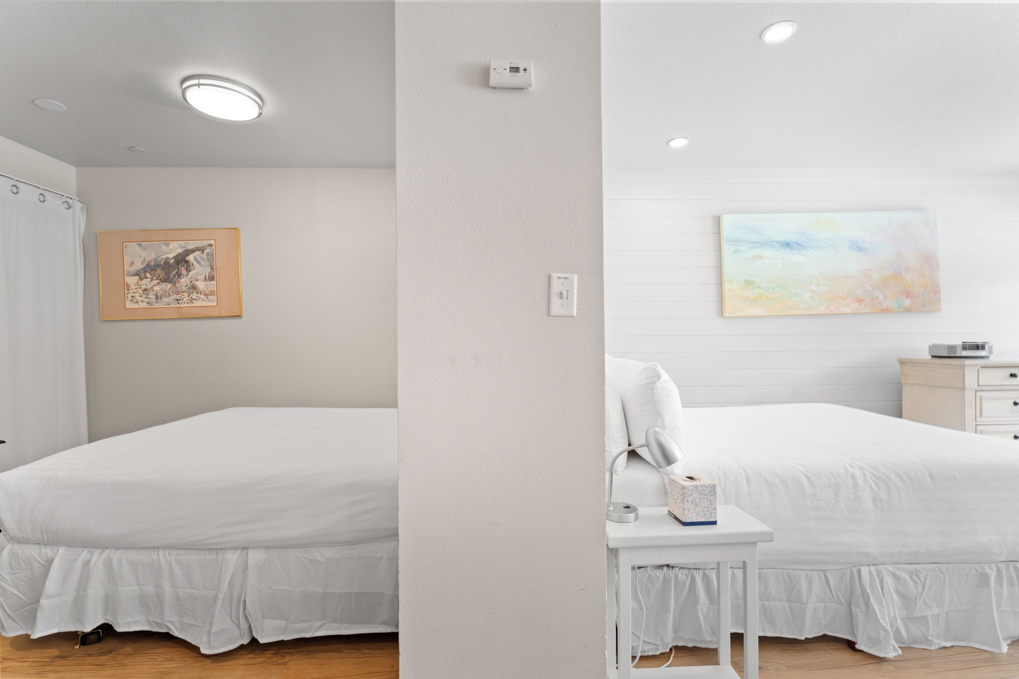 A bedroom with two beds, a nightstand, a lamp, a painting, and a thermostat on the wall.