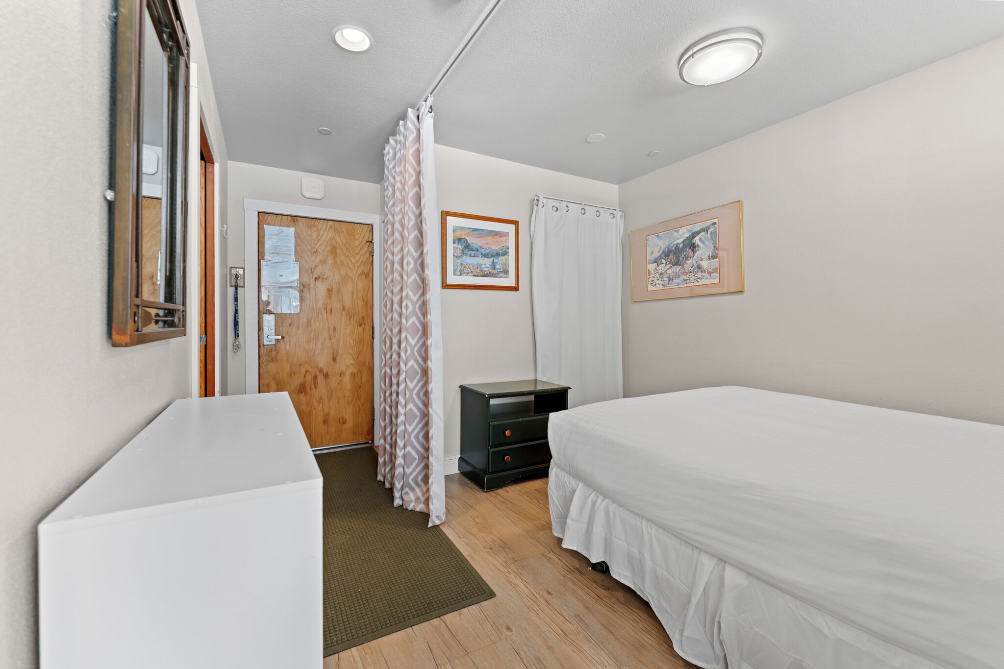 Un dormitorio con una cama, una mesita de noche, una puerta de madera, un espejo y dos cuadros en la pared.