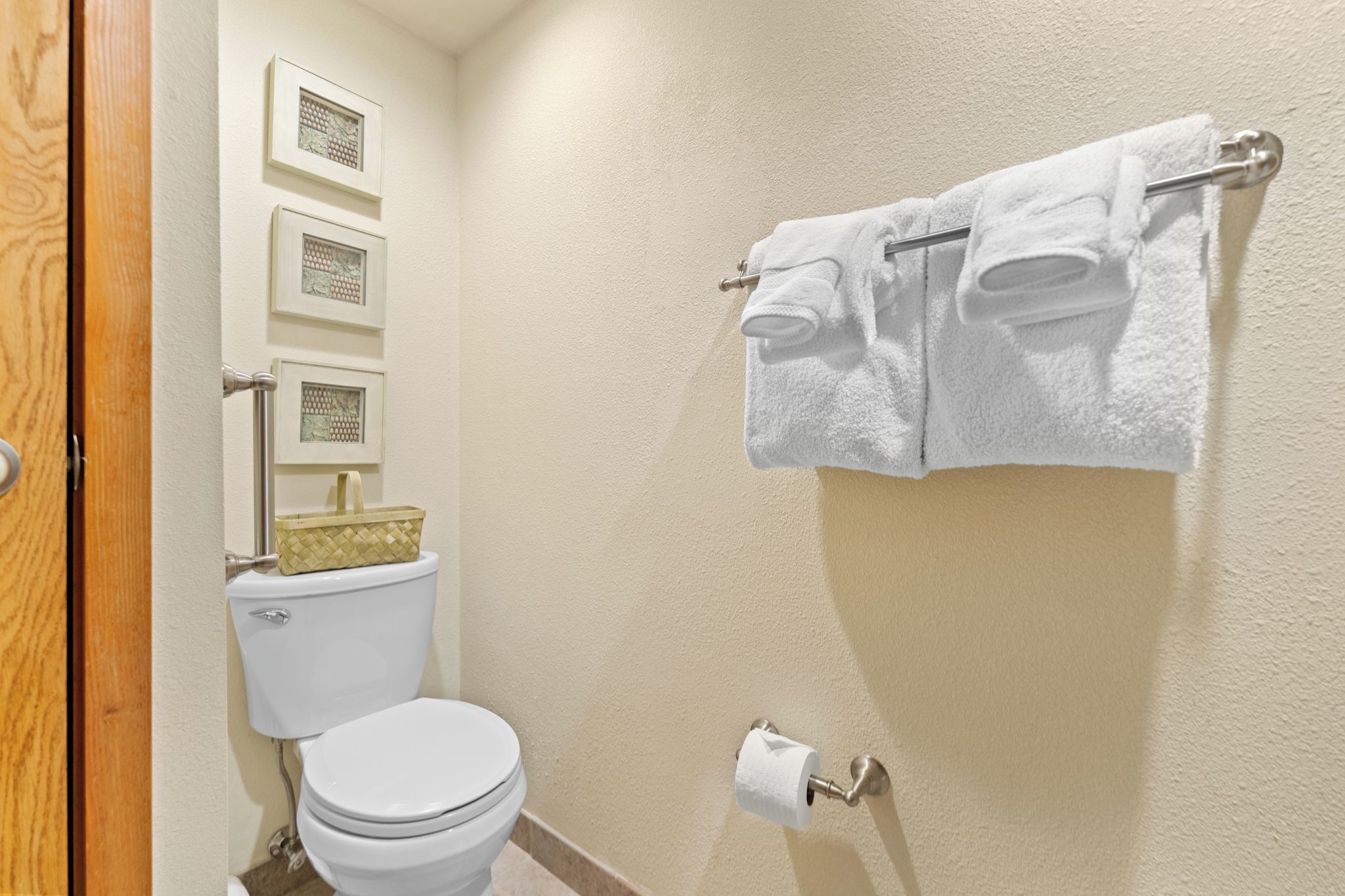 A bathroom with a toilet, a towel rack with two towels, a toilet paper holder, and three framed pictures on the wall.