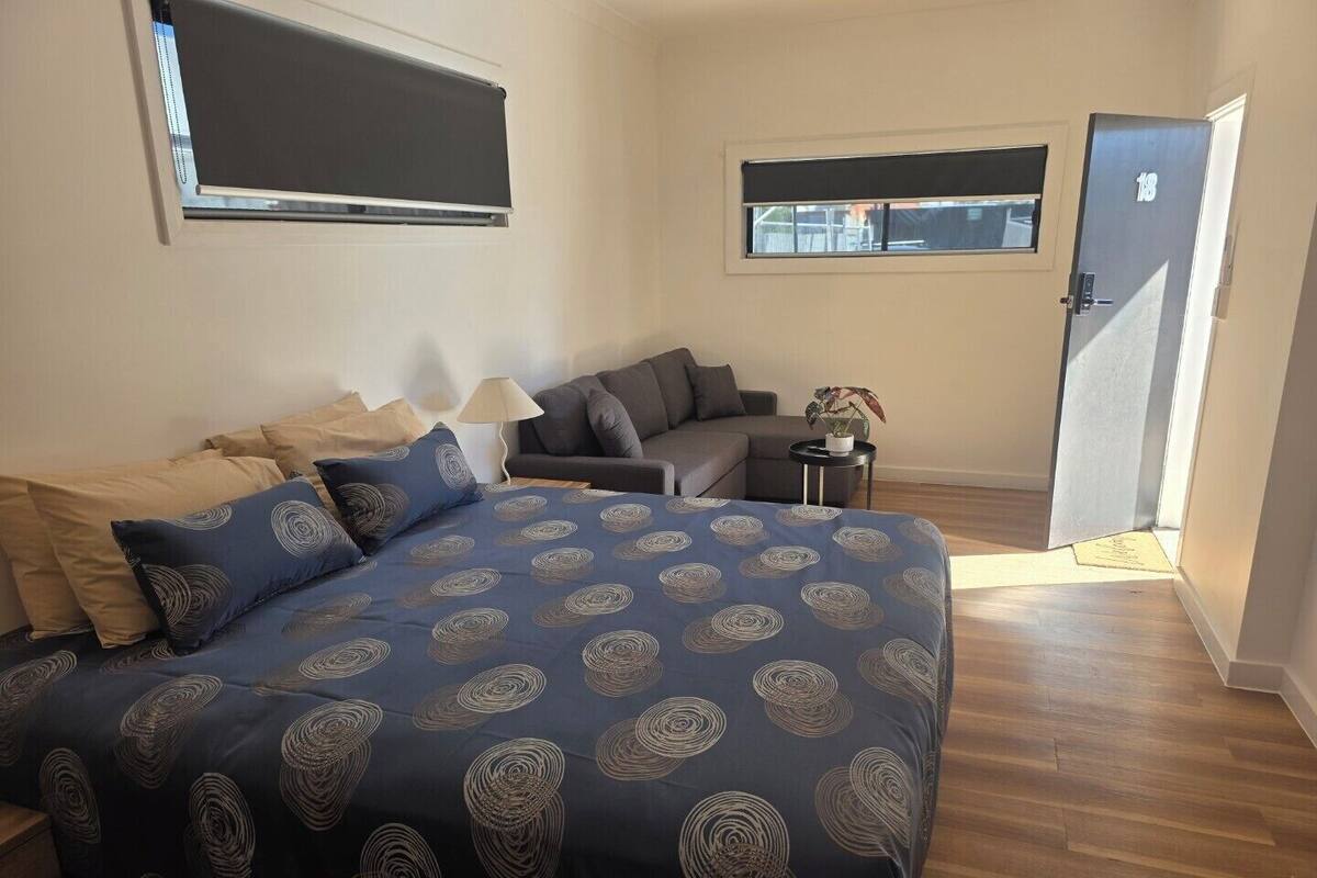 A bedroom with a bed, a sofa, a small table with a lamp, and a door.
