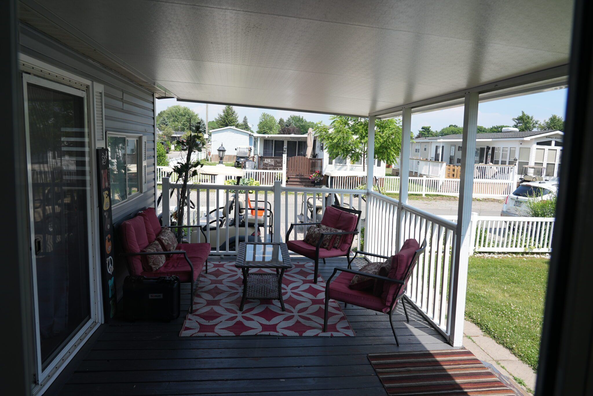 A covered porch with a table and chairs, a white picket fence, and a view of mobile homes in the background.