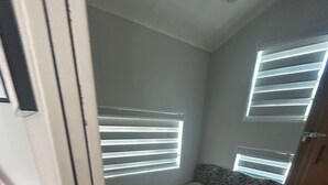 Um quarto pequeno e estreito com uma cama de solteiro, uma janela com persianas e uma porta.