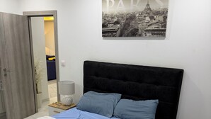 Een slaapkamer met een bed, nachtkastje, lamp, een deur en een aan de muur hangend schilderij van Parijs.