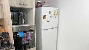 Een keuken met een koelkast, oven en magnetron. Er staat een tafel met een schaal fruit en een fles.
