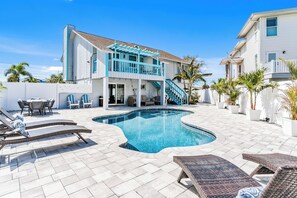 Ein Schwimmbecken mit Liegestühlen, einer überdachten Terrasse und einem zweistöckigen Gebäude mit blauer Brüstung.