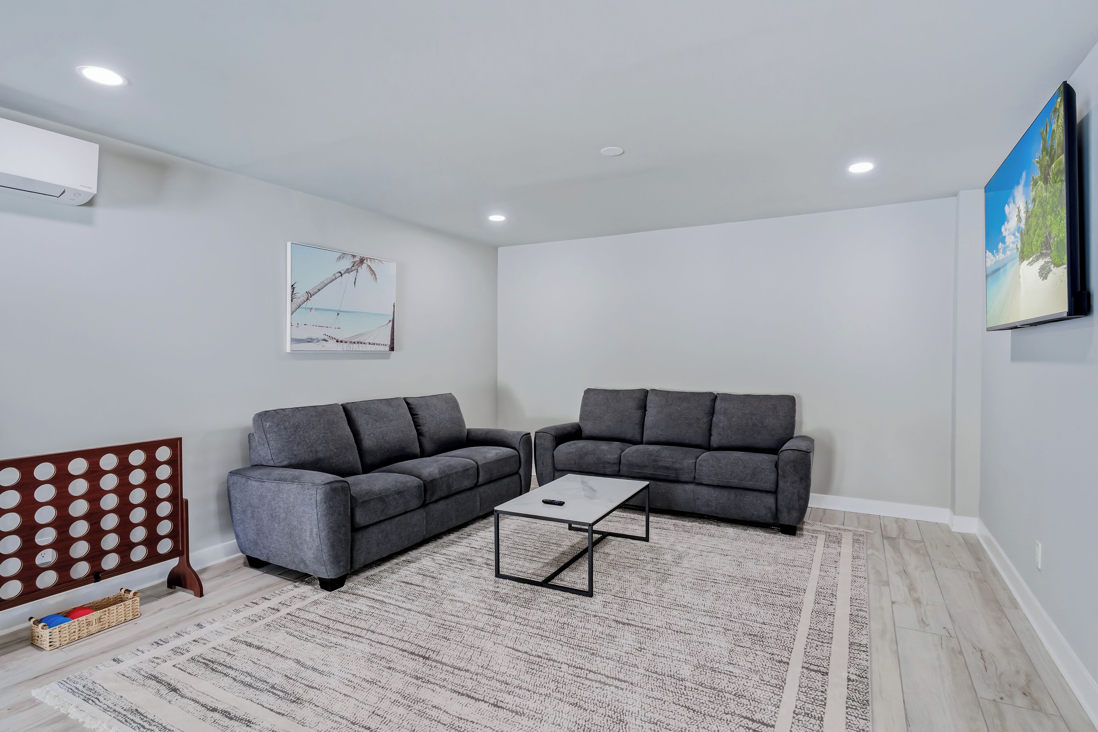 Un salon avec deux canapés gris, une table basse, un tapis à motifs, un téléviseur fixé au mur et un tableau représentant une scène de plage.