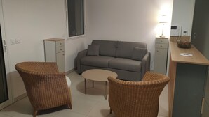 Un petit salon comprenant un canapé, deux fauteuils en osier, une petite table, une fenêtre et un coin cuisine.