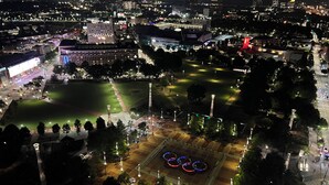 Vista aérea de un paisaje urbano nocturno con edificios iluminados, una amplia plaza abierta de diseño circular y una estructura roja destacada.