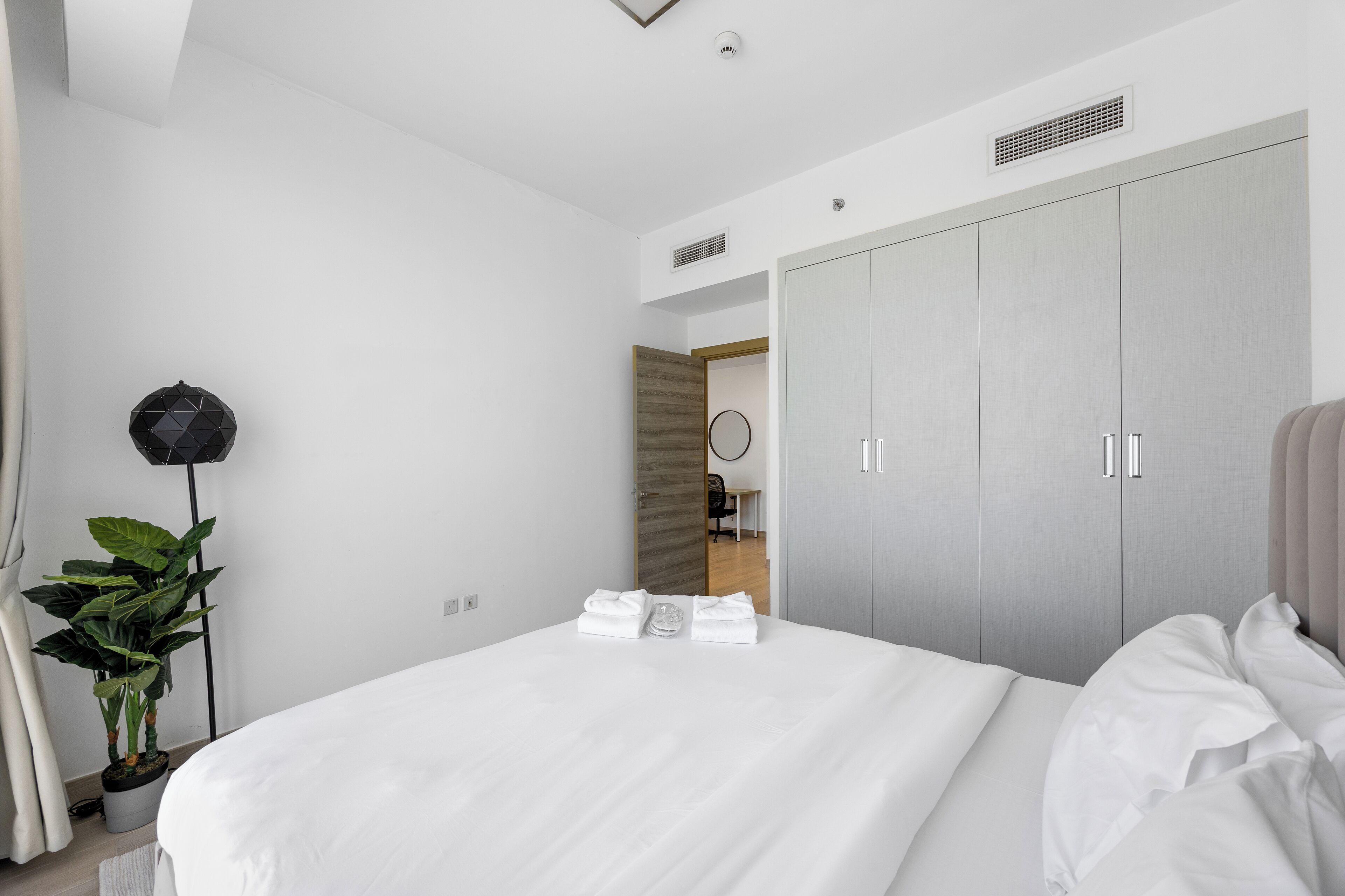 A modern hotel room with a large bed, grey wardrobe, potted plant, and a ceiling light fixture.