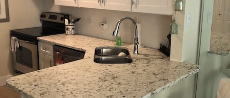 Une cuisine avec des meubles blancs, un plan de travail en granit, un évier, une cuisinière et deux tabourets de bar.
