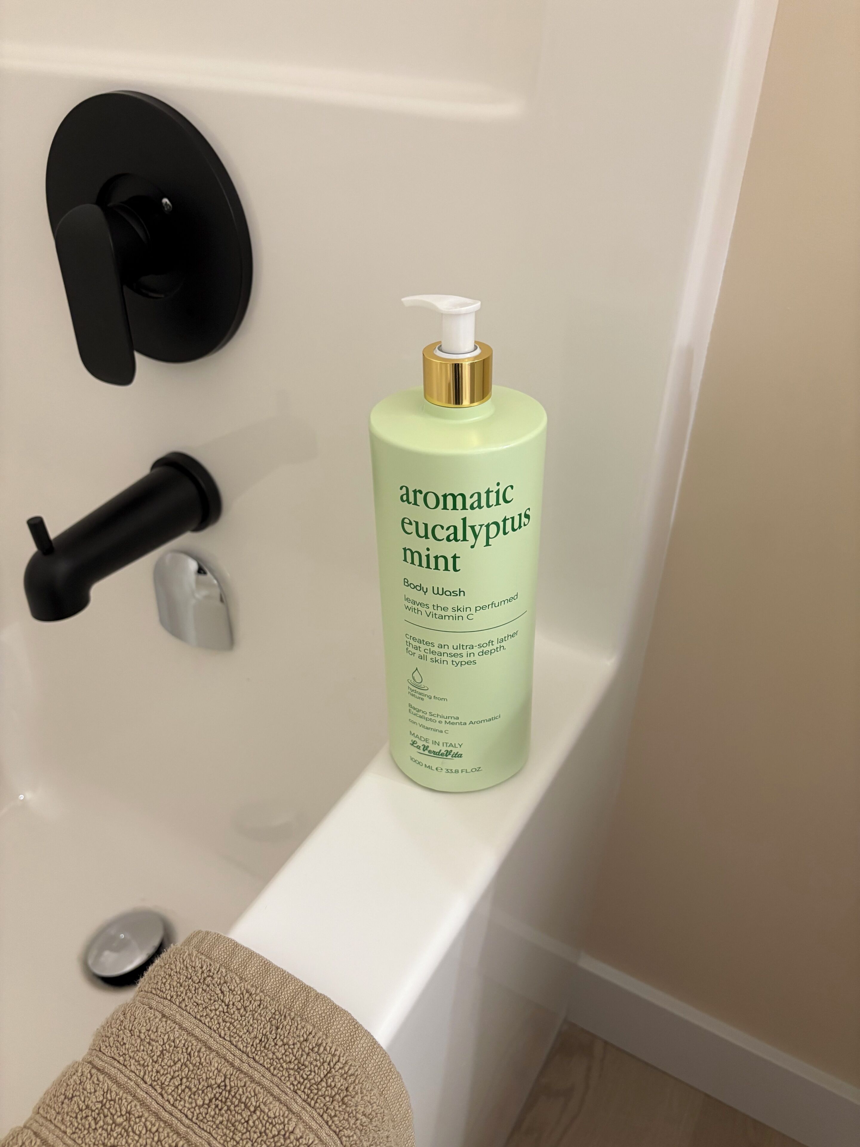 Baño con una bañera blanca, una botella de jabón corporal aromático de eucalipto y menta, una toalla negra y un objeto negro en la pared.