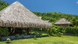 Uma construção com telhado de palha, varanda e cadeiras, rodeada por vegetação e um céu claro.