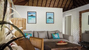 Uma sala de estar com uma mesa de centro de madeira, um sofá com almofadas, uma poltrona de vime e dois quadros decorativos na parede.