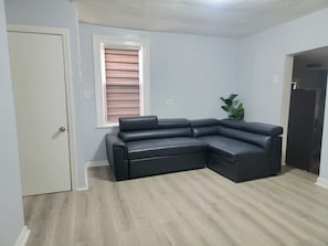 A room with a sectional sofa, a door, a window with blinds, a plant, and a dark-colored cabinet.