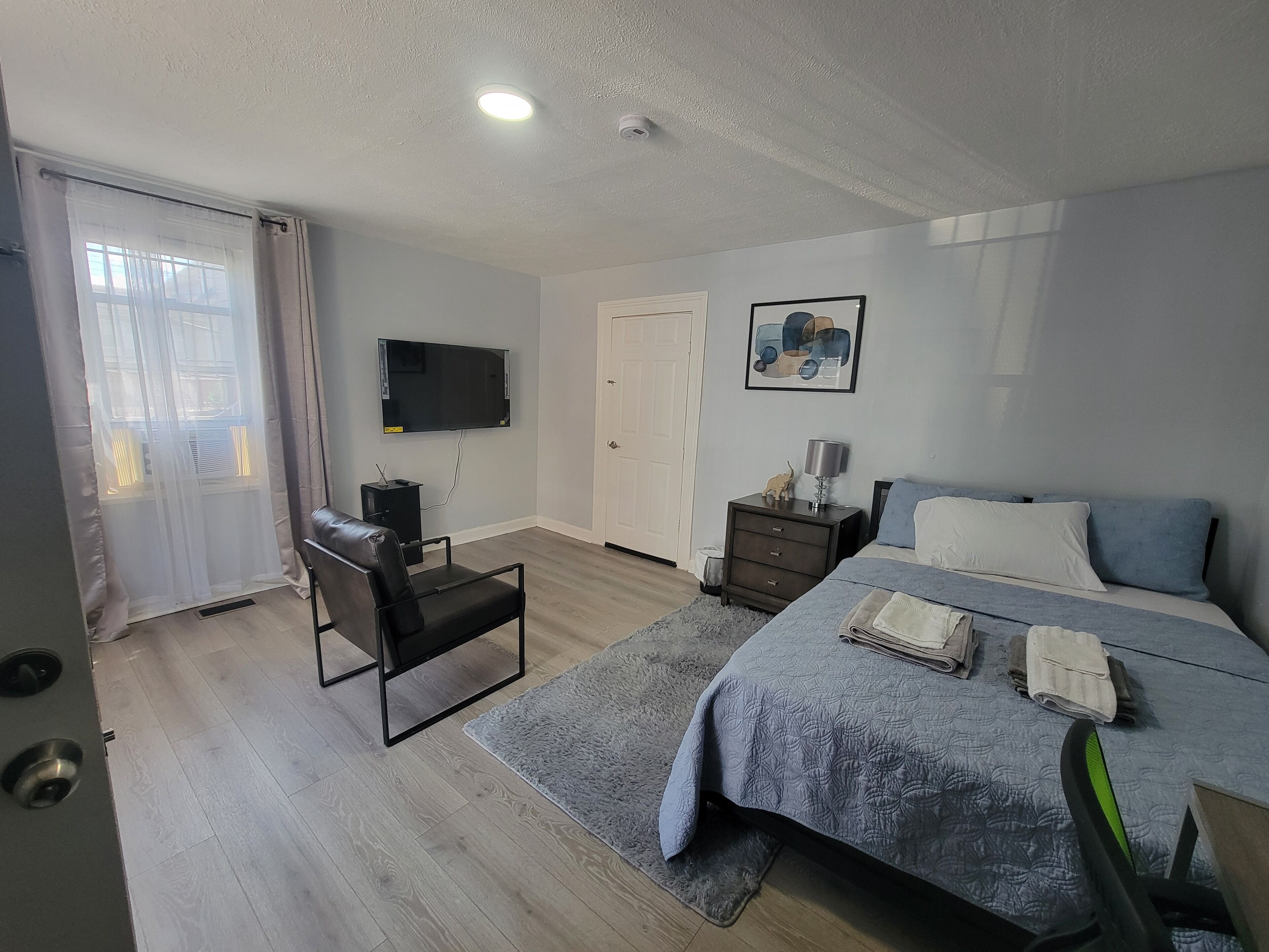 A bedroom with a bed, a desk, a chair, a TV, a door, and a small artwork on the wall.
