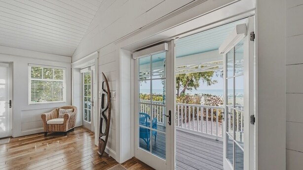 A room with wooden flooring, a wicker chair, a large glass door leading to a balcony with a view of the beach. - Happy Ours – NEW LISTING, Direct Beachfront Home On Magnolia (Anna Maria)