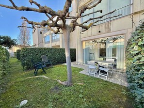 Une cour bordée d’arbres, dotée d’un espace extérieur aménagé avec des sièges, et un bâtiment aux grandes fenêtres.