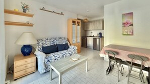 En kompakt opholdsstue med sofa, bord, lampe, tekøkken og spiseplads.