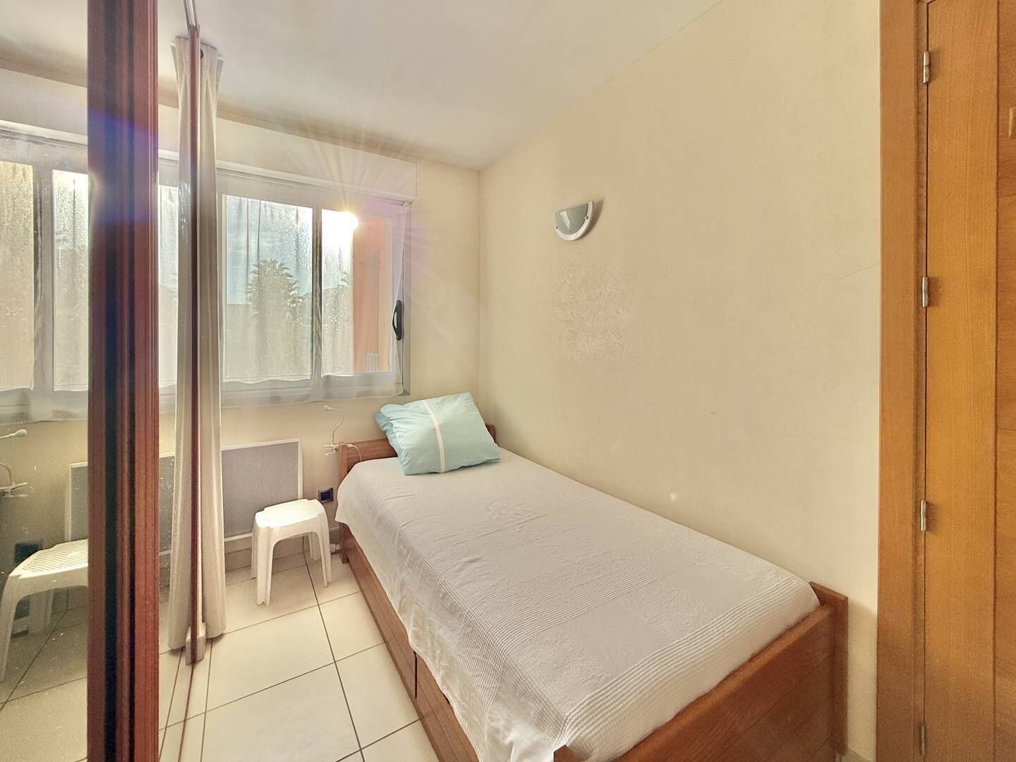 A bedroom with a bed, a small stool, a window, and a wooden door.