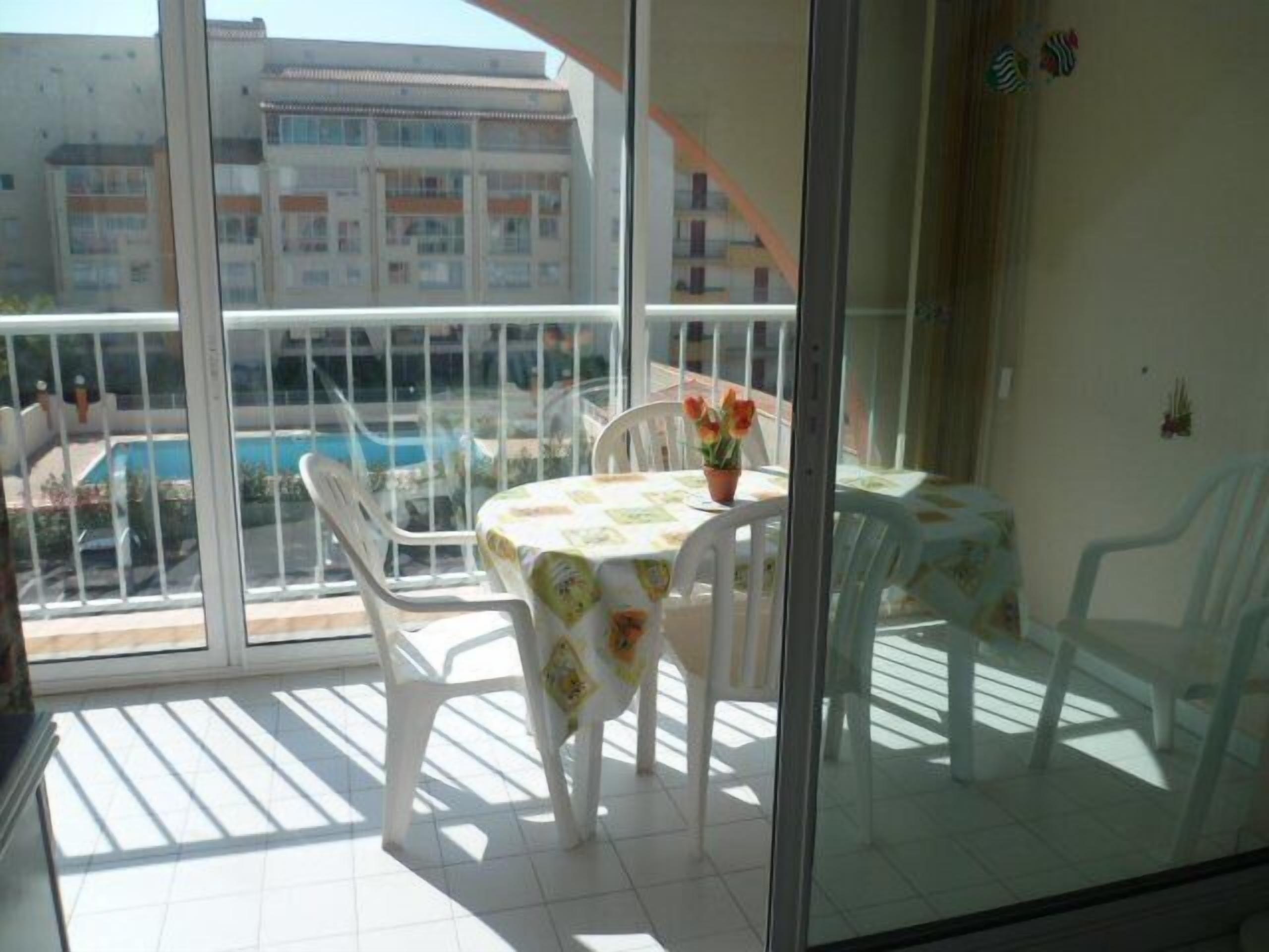 Un balcon avec une table et des chaises, un pot de fleurs et une vue sur un immeuble et une piscine.