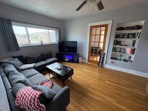 Ein Wohnzimmer mit Ecksofa, Couchtisch, Flachbildfernseher, Bücherregal und einer Tür, die nach draußen führt.