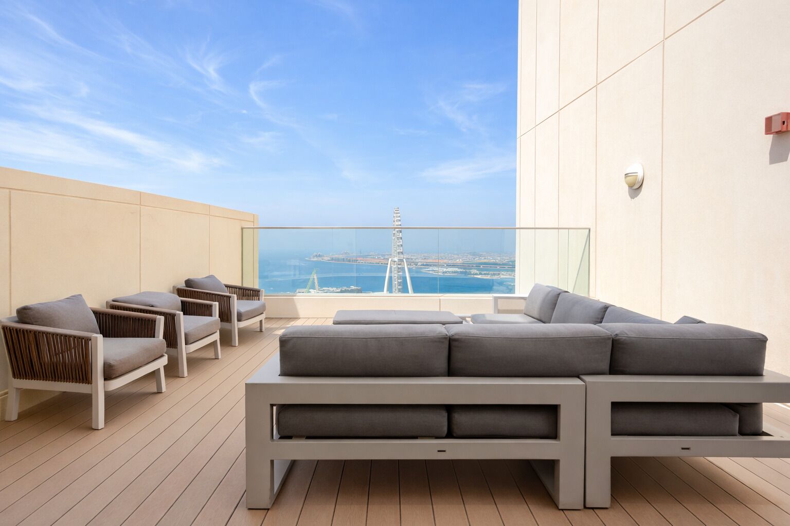 A modern outdoor seating area with a sofa and chairs, a glass balustrade, and a view of a distant cityscape and water.