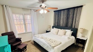 Une chambre à coucher comprenant un lit, un ventilateur de plafond, une fenêtre avec des rideaux, une table de chevet et une chaise.