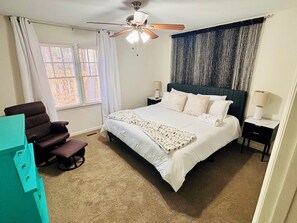 A bedroom with a bed, a ceiling fan, a window with curtains, a nightstand, and a chair. - Walk to Shoppes of Kildaire & Close to Bond Park! (Cary)